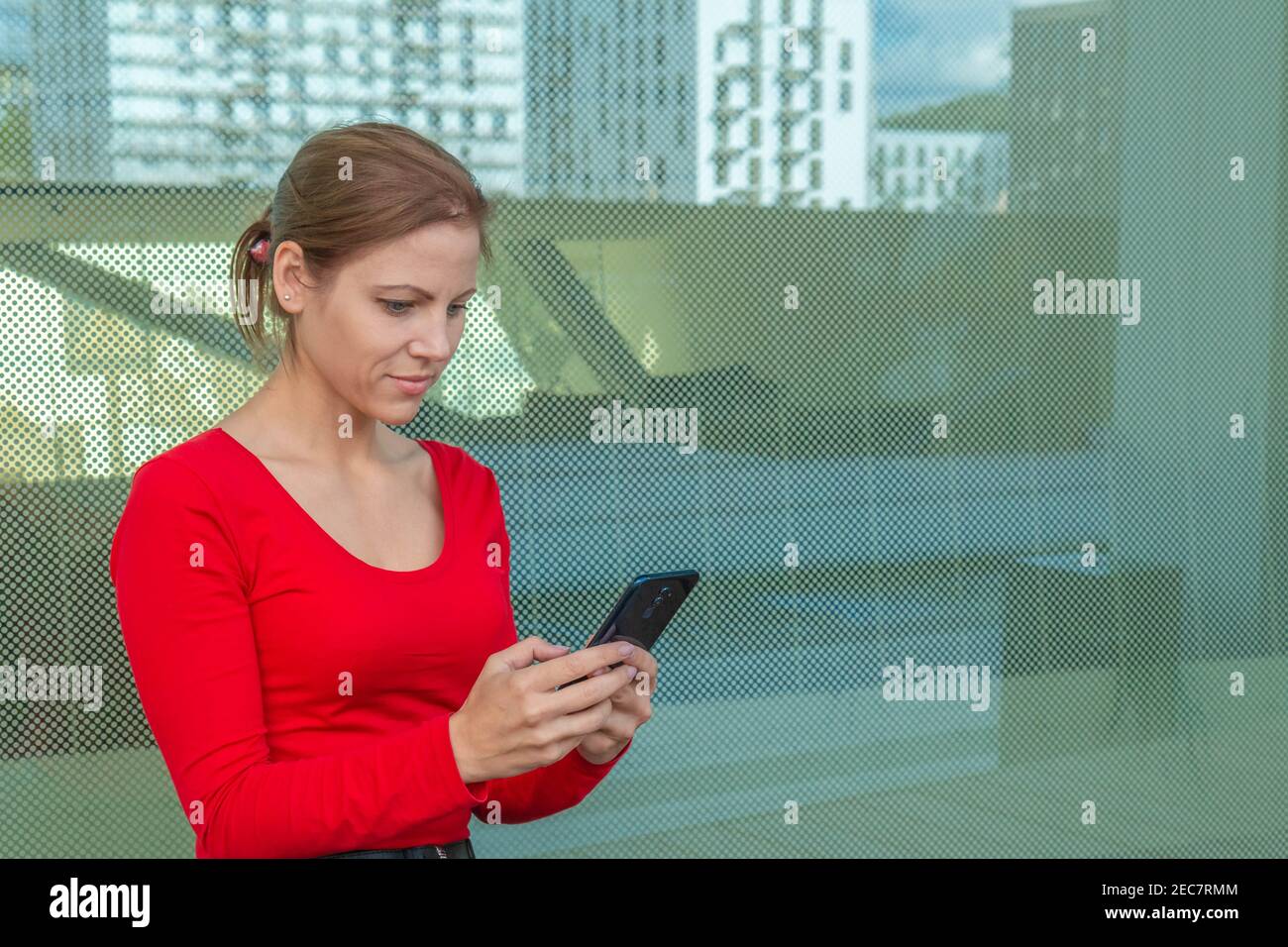 Belle femme en rouge debout dans le mur de verre avant avec réflexion et en utilisant le smartphone. Copier l'espace sur le reflet de la vue du bâtiment moderne. Banque D'Images