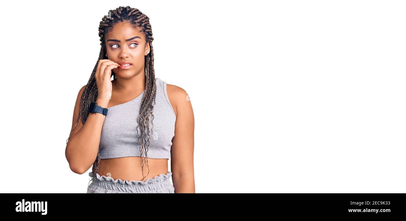 Jeune femme afro-américaine avec des braïdes portant des vêtements de sport semble stressée et nerveuse avec les mains sur la bouche piquant les ongles. Problème d'anxiété. Banque D'Images