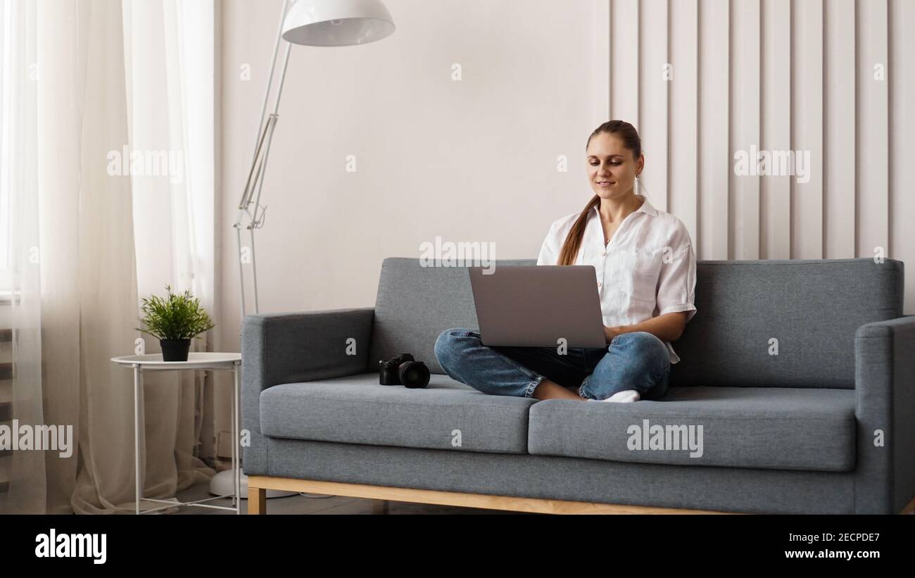 Bonne jeune femme utilisant un ordinateur portable à la maison. Sur le canapé à côté de la femme se trouve une caméra. Le photographe retouche des photos à la maison. Banque D'Images