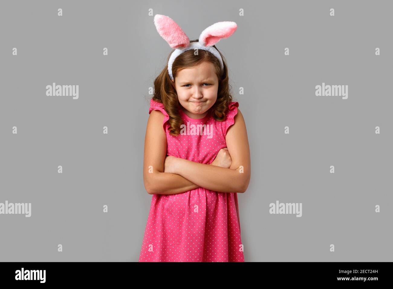 Petite fille dans une robe rose avec des pois sur un fond gris. Enfant dans les oreilles de lapin de Pâques en colère avec les bras croisés. Banque D'Images