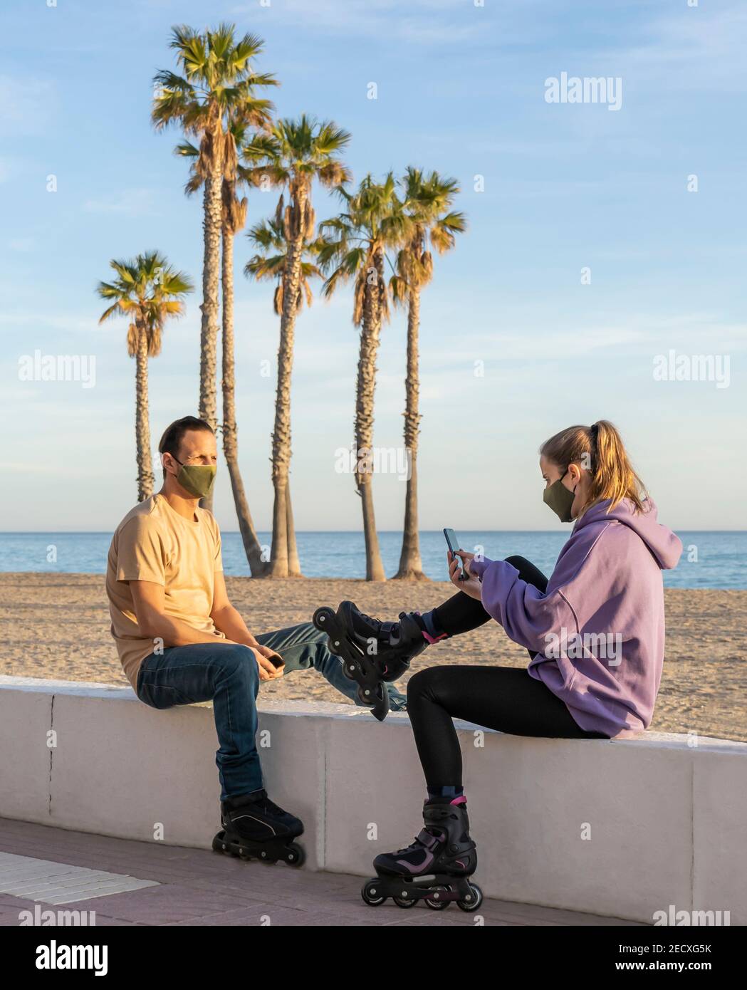 Couple de patinage à roulettes dans un masque médical de protection (contre le coronavirus , Covid-19) assis sur la promenade, en utilisant le téléphone cellulaire sur le bord de mer, les palmiers et le beac Banque D'Images