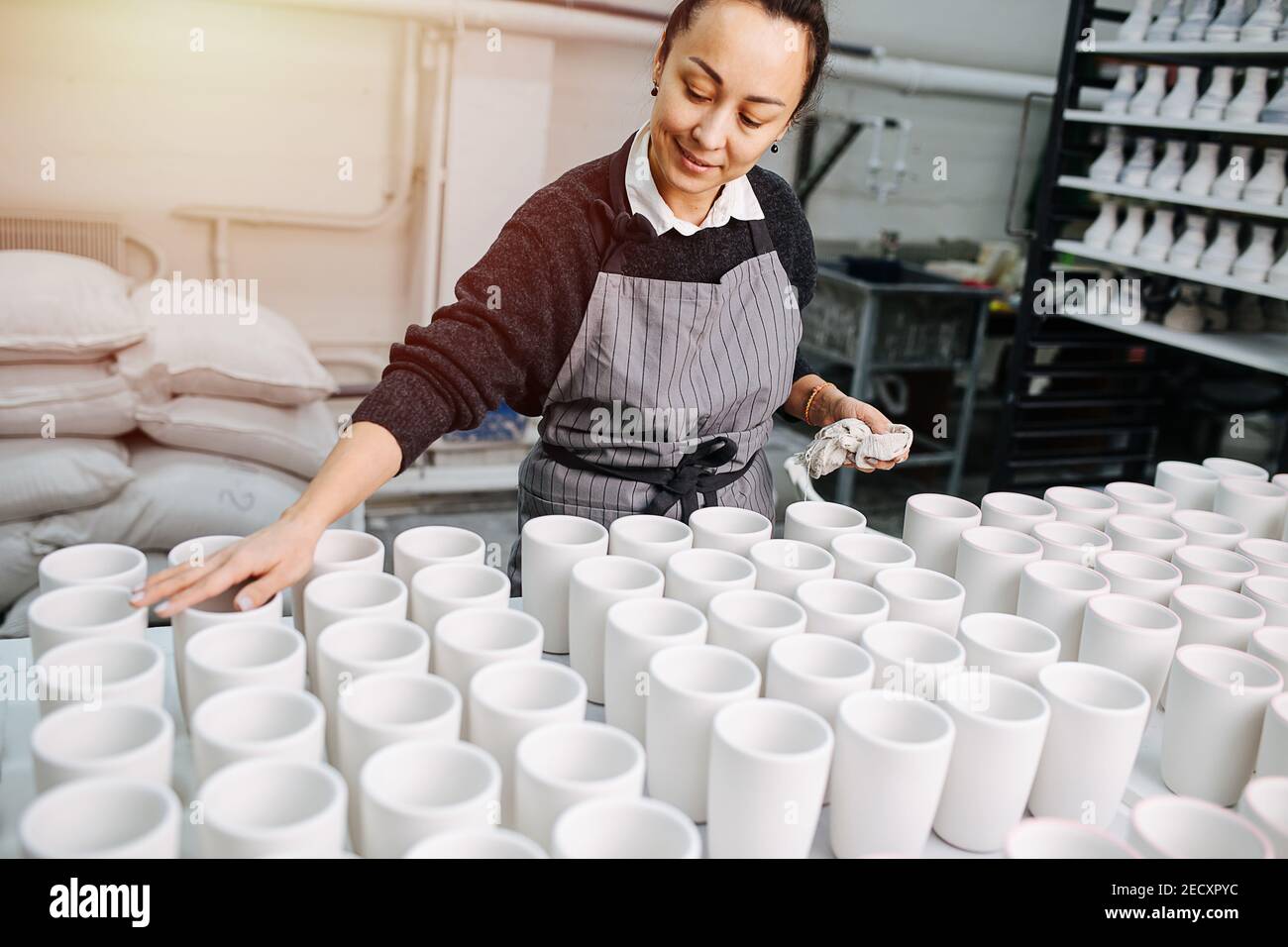 Femme fière de nettoyer des rangées de tasses vierges, travaillant dans un entrepôt de poterie Banque D'Images