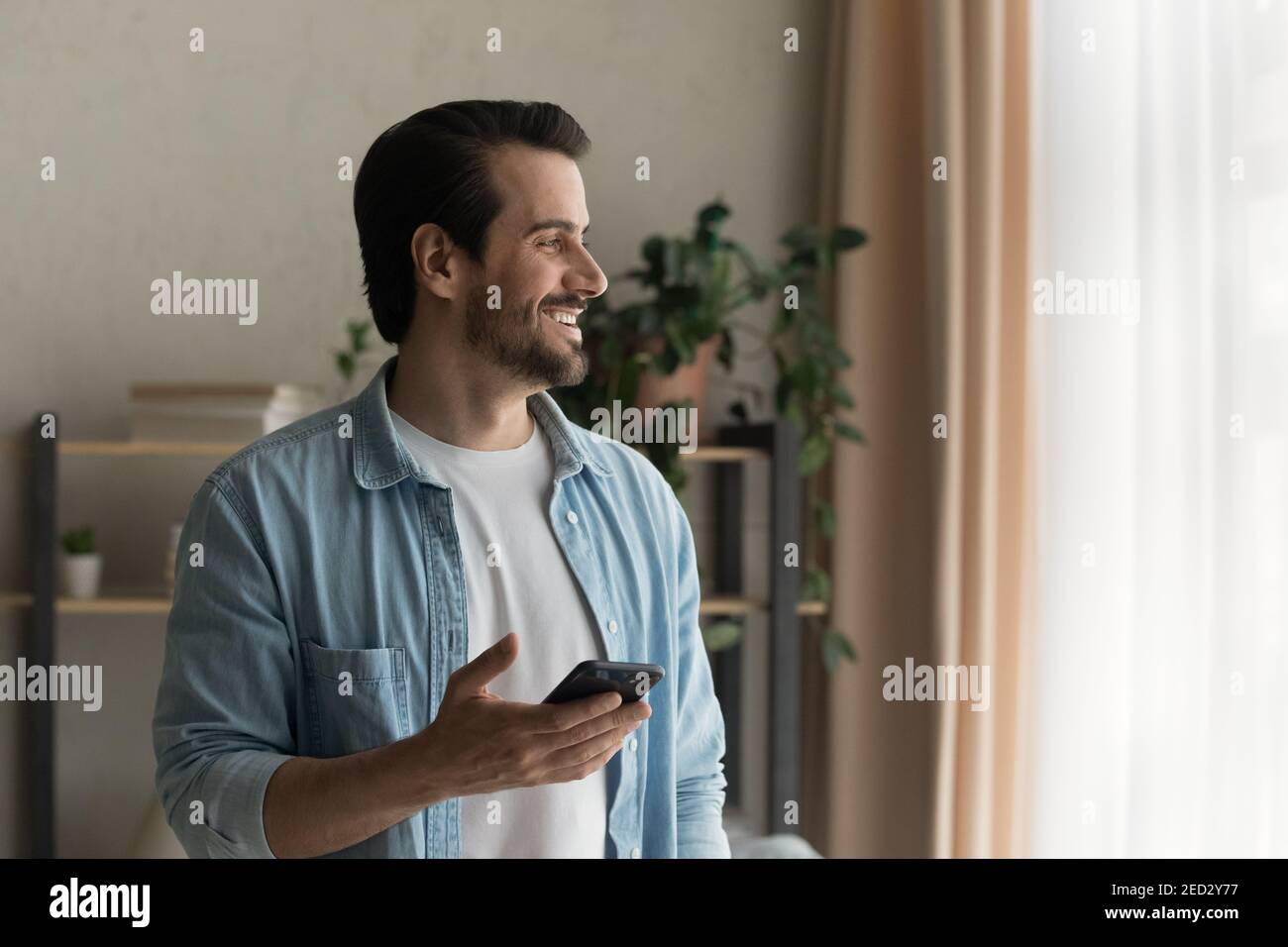 Un homme heureux, debout près de la fenêtre, ne regarde pas distrait du smartphone Banque D'Images