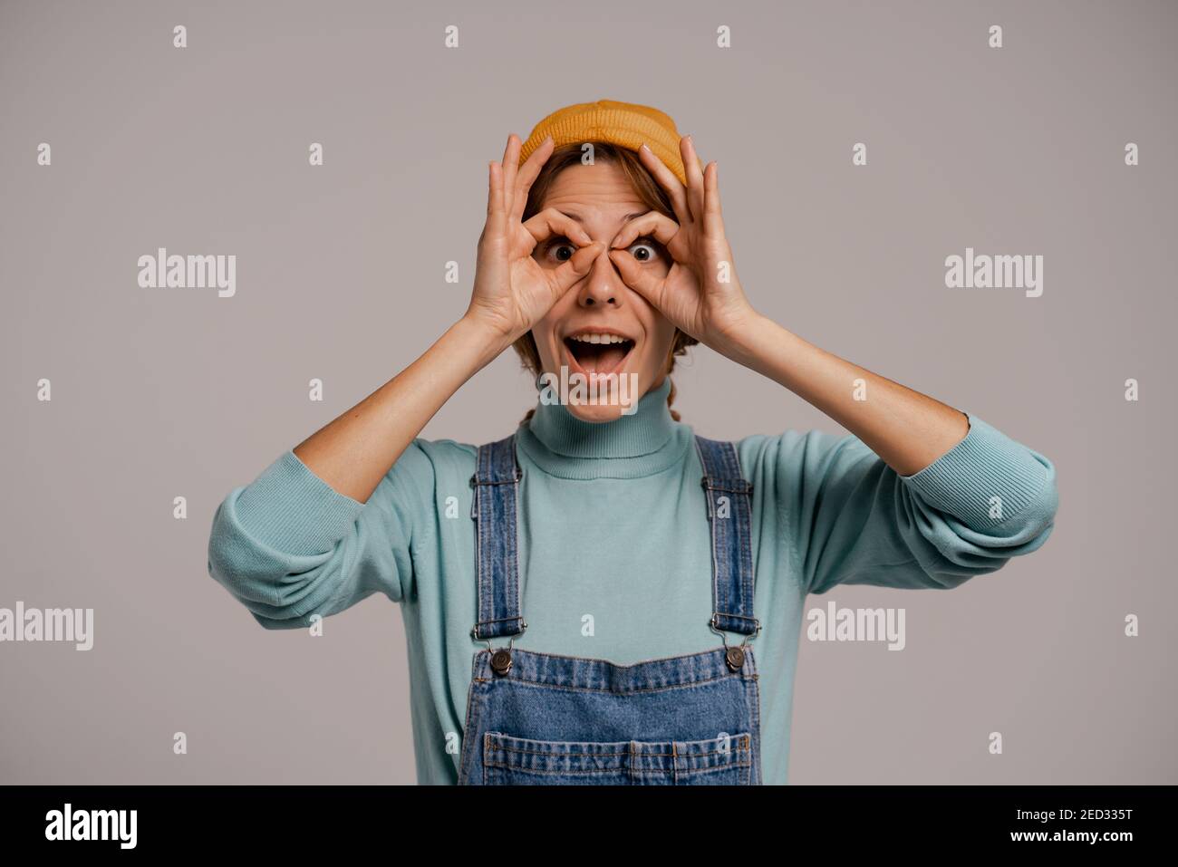 Photo de la jolie femelle hipster tend à regarder dans les verres. Blanc femme porte une combinaison en denim et chapeau isolé de couleur grise fond Banque D'Images