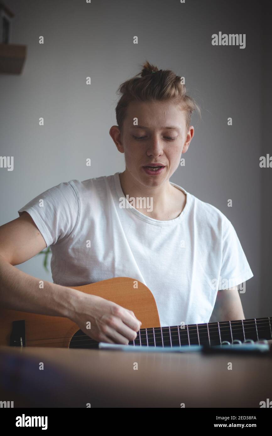 un garçon talentueux jouant une guitare acoustique en bois dans une salle éclairée. Homme blond âgé de 20-29 ans, guitare et chant d'un livre d'hymne sur un canapé. Instrument de musique Banque D'Images