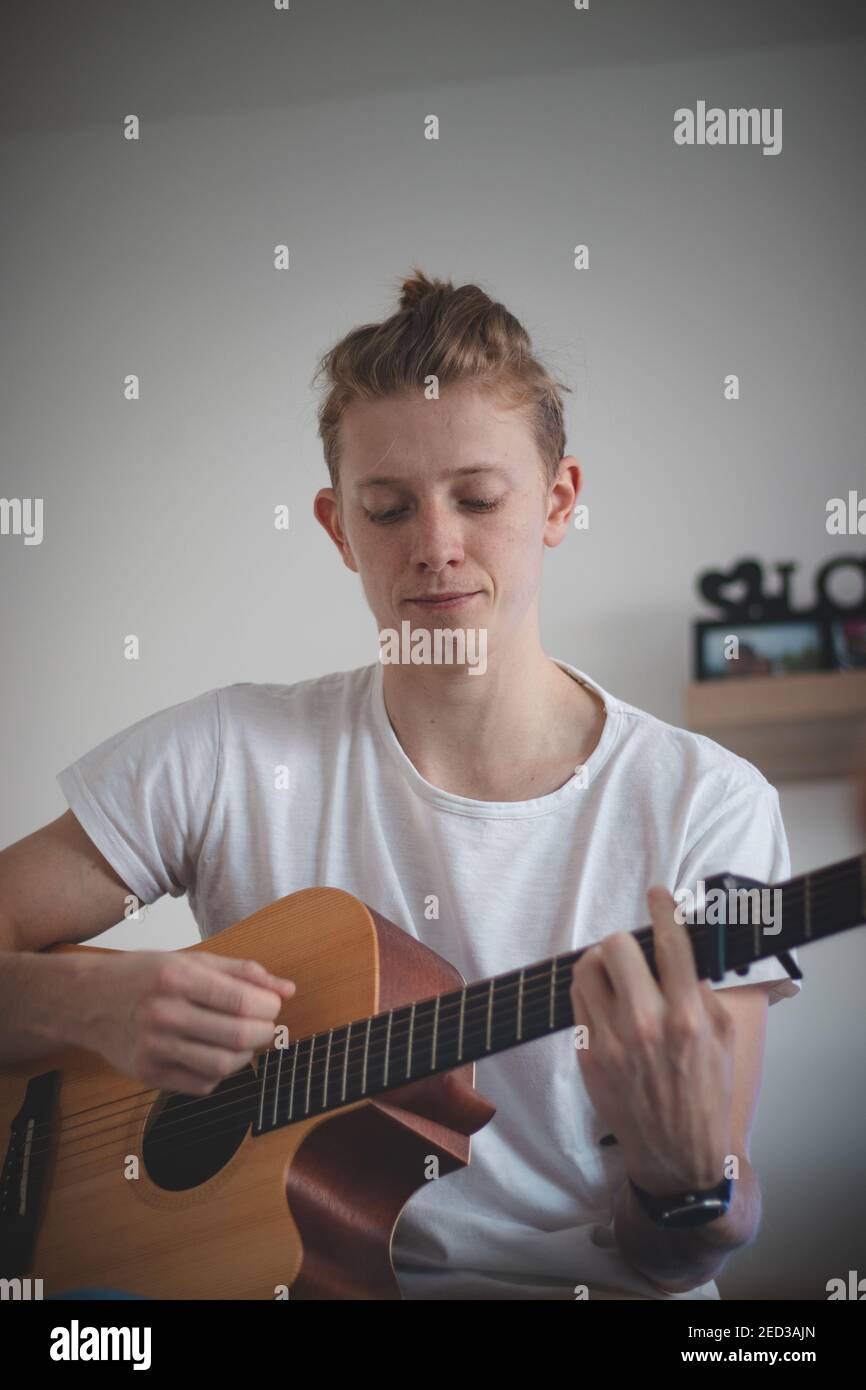 un garçon talentueux jouant une guitare acoustique en bois dans une salle éclairée. Homme blond âgé de 20-29 ans, guitare et chant d'un livre d'hymne sur un canapé. Instrument de musique Banque D'Images