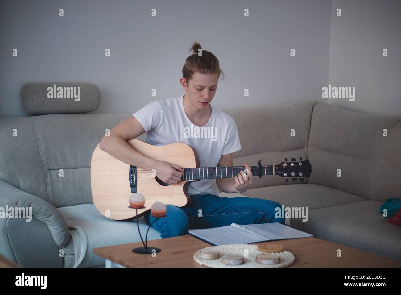 un garçon talentueux jouant une guitare acoustique en bois dans une salle éclairée. Homme blond âgé de 20-29 ans, guitare et chant d'un livre d'hymne sur un canapé. Instrument de musique Banque D'Images