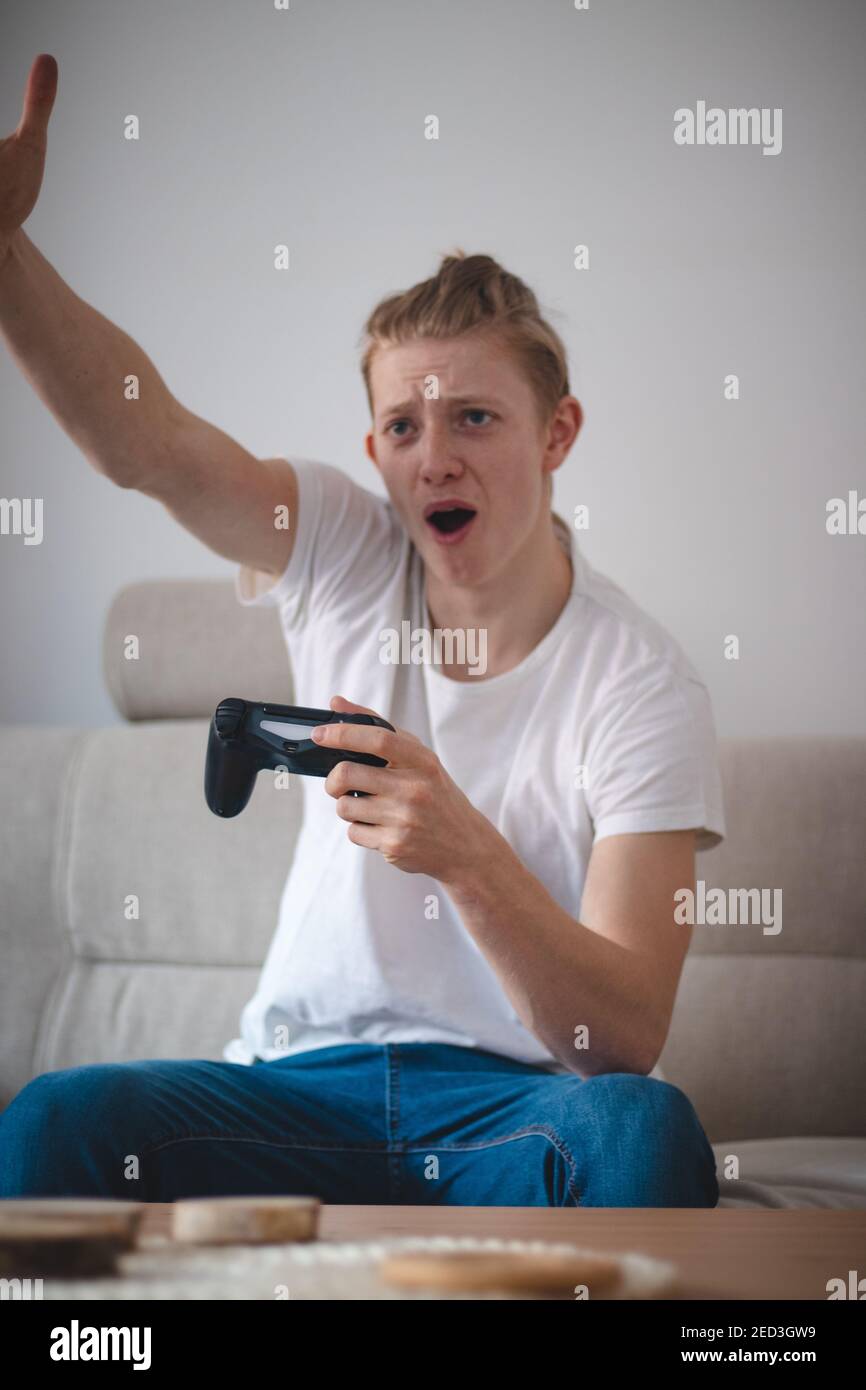 Un jeune homme en colère assis sur un canapé dans sa salle de séjour moderne qui jette les bras tout en jouant à un jeu de stratégie à la télévision. D'une main, il tient le contrôle Banque D'Images