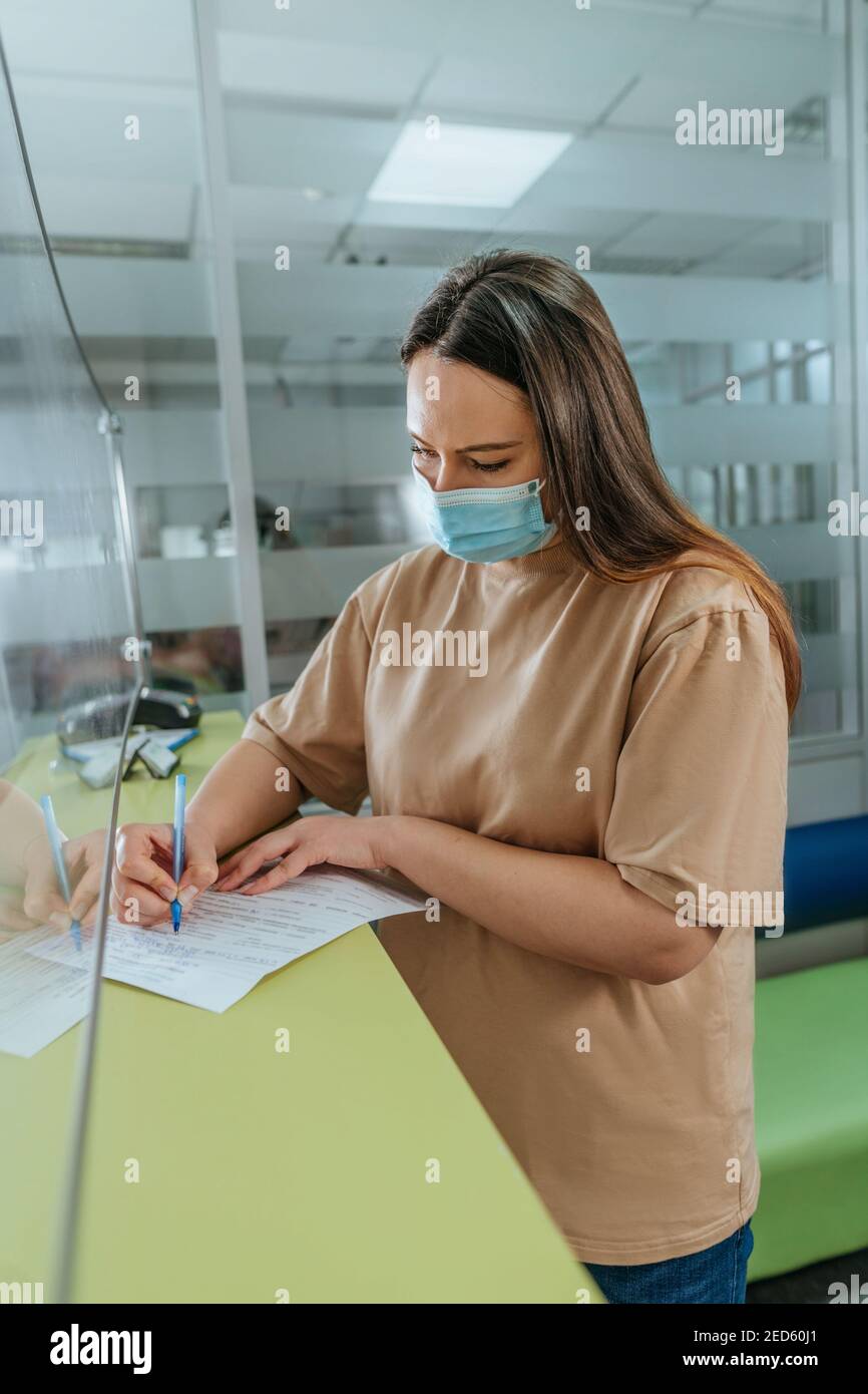 Femme dans le masque facial protecteur remplissant la forme médicale à réception de l'hôpital au comptoir dans le hall avant un examen médical Banque D'Images