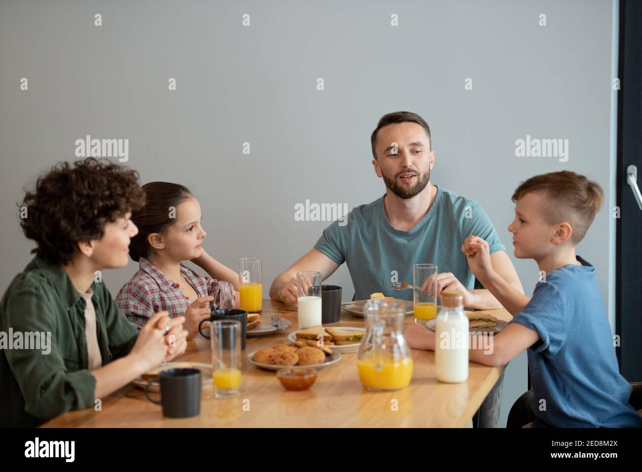 Bonne famille de quatre personnes prenant le petit déjeuner servi à table dans la cuisine tout en regardant le petit garçon et en écoutant à ses idées pour le week-end Banque D'Images