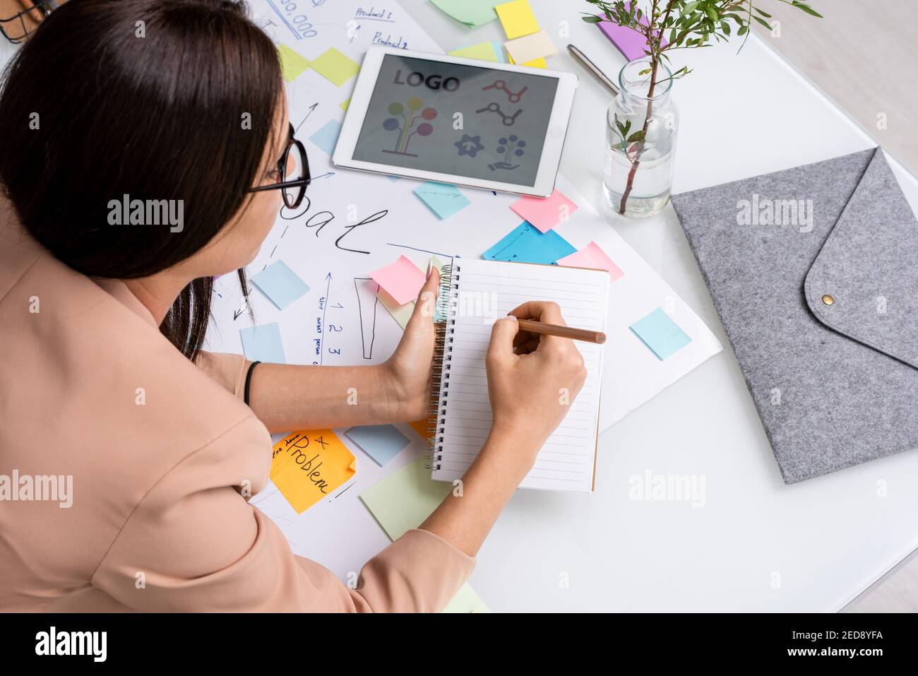 Jeune femme d'affaires brunette se pliant sur un document de grande taille avec des objectifs principaux et des points de travail lors de la prise de notes sur la page de l'ordinateur portable Banque D'Images