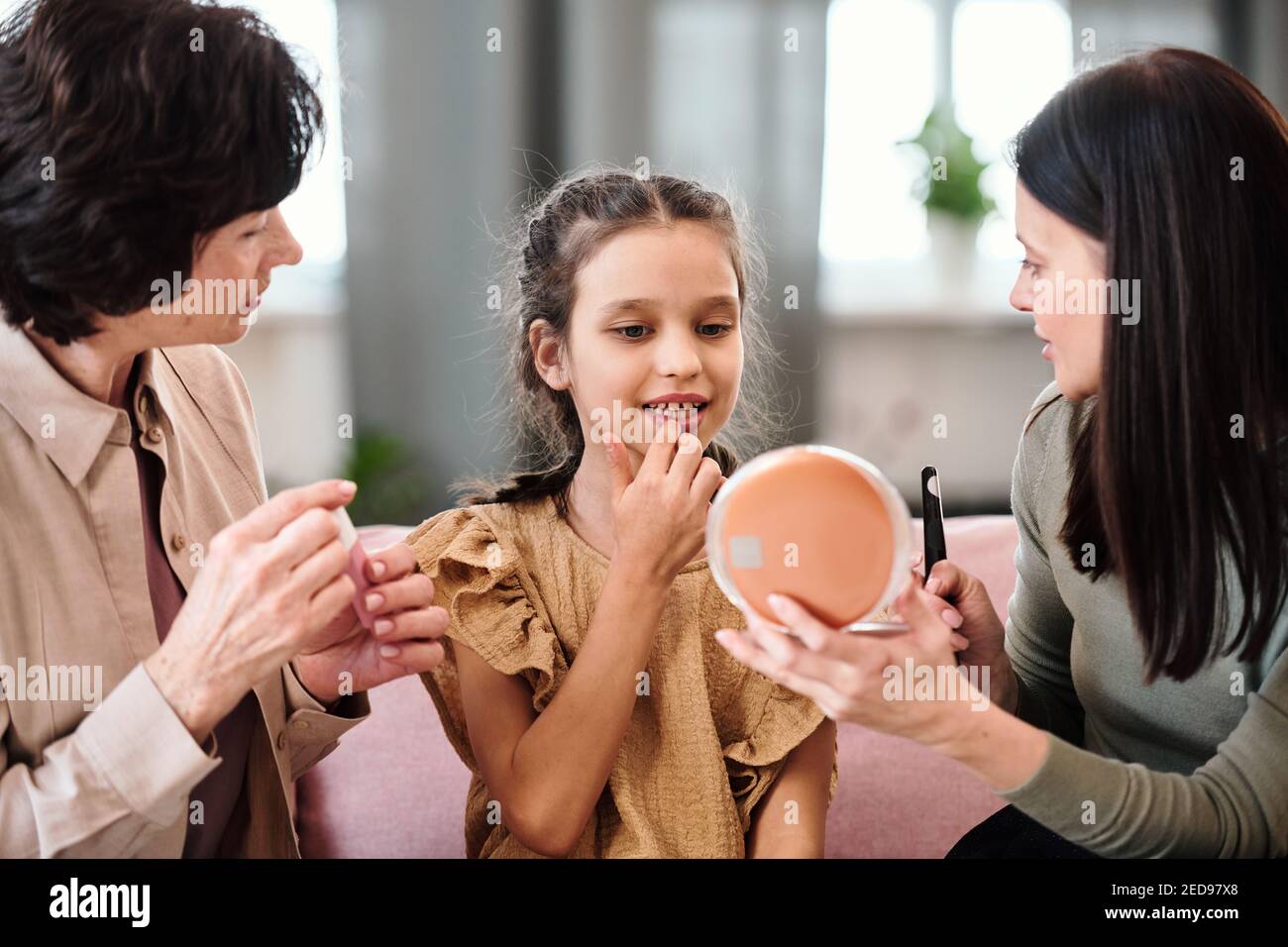 Jolie petite fille en robe appliquant un baume nutritif sur les lèvres tout en regardant dans le miroir tenu par la jeune femme brunette donnant ses conseils de beauté Banque D'Images