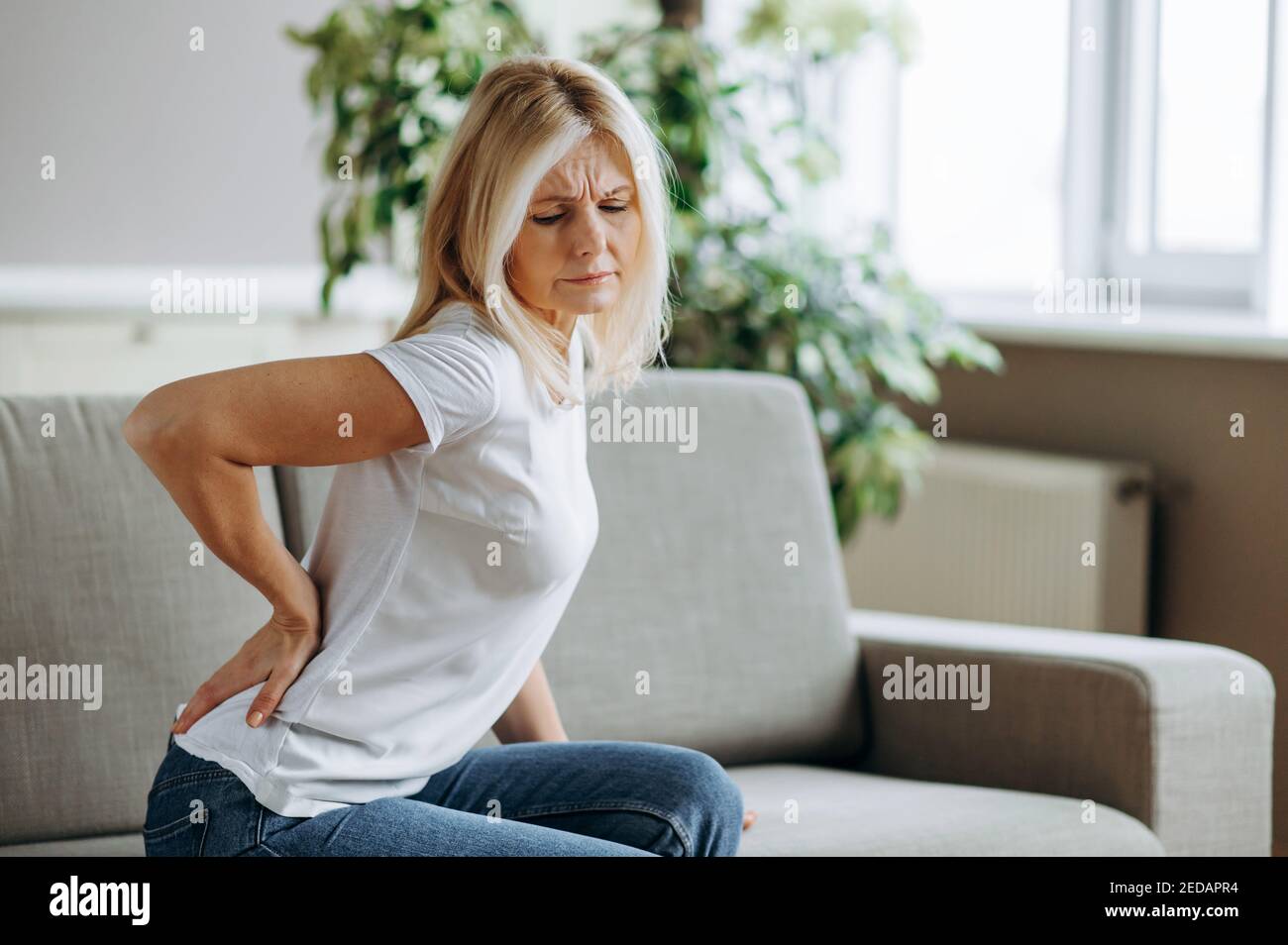 Contrarié femme mature souffrant de mal de dos, blond senior malheureux assis sur un canapé dans le salon, sensation d'inconfort à cause de la douleur dans le dos. Banque D'Images