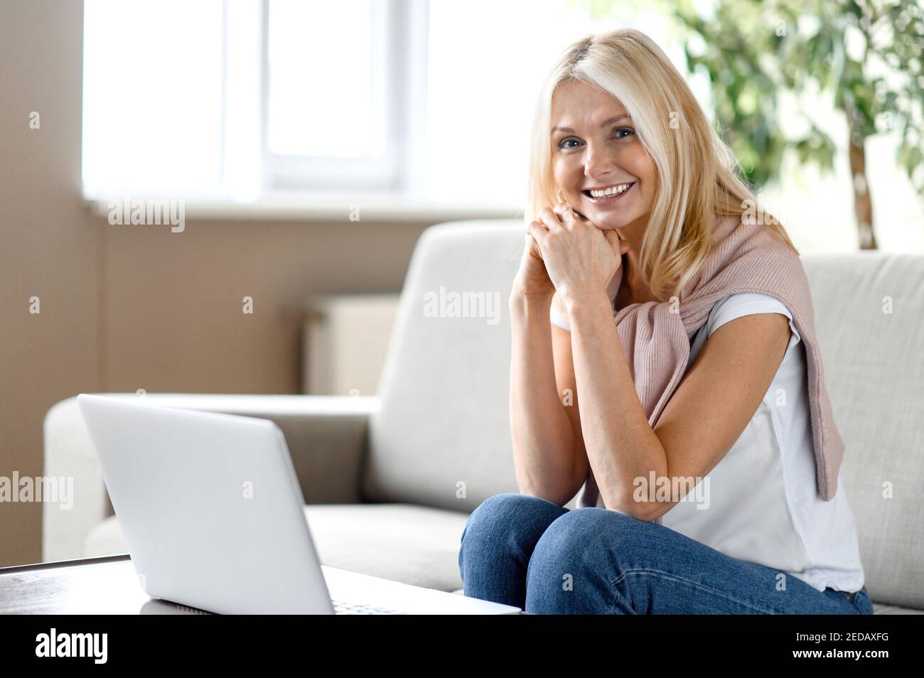 Décontractée femme blonde de race blanche d'âge moyen assise à la maison dans le salon, à l'aide d'un ordinateur portable, regarde l'appareil photo et sourire heureux, travailler ou étudier à distance Banque D'Images