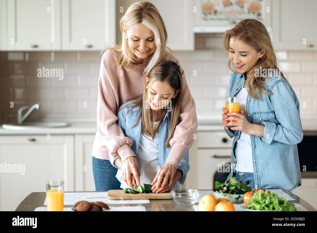 Trois générations de femmes passent du temps ensemble dans la cuisine, la grand-mère enseigne à la petite-fille de couper la salade et les montres de maman souriant et de boire du jus à côté d'eux Banque D'Images