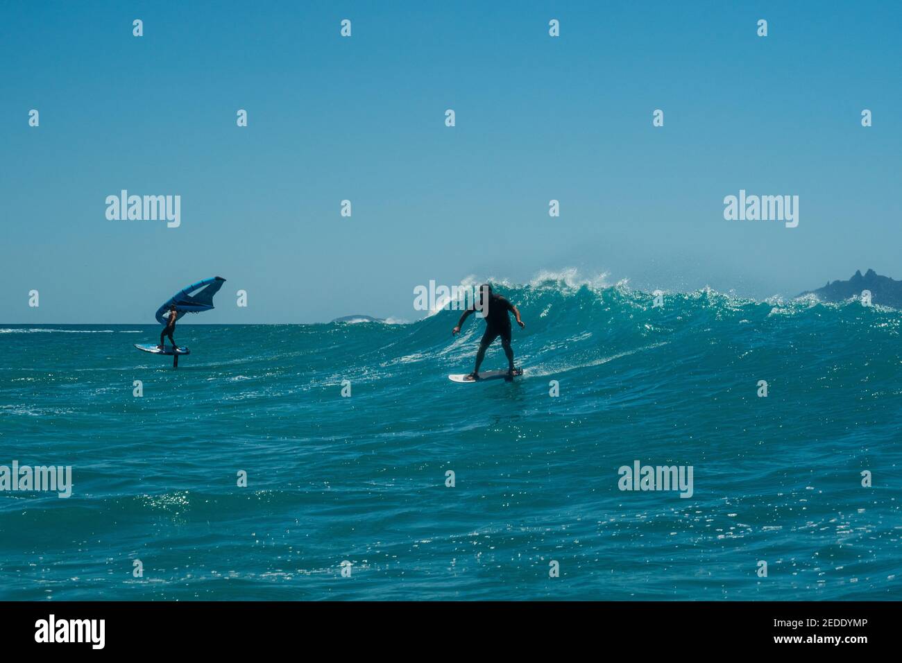 Deux hommes surfent sur une vague à l'aide de planches de surf hydrofoil, un homme à l'aide d'une aile à la main. Bleu turquoise mer, ciel bleu clair. Banque D'Images