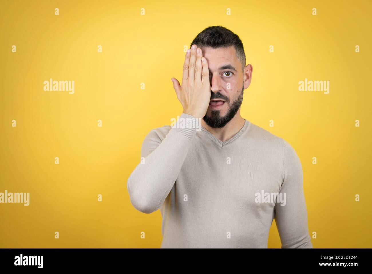 Beau homme avec une barbe portant un pull sur fond jaune couvrant un œil avec la main, un sourire assuré sur le visage et une émotion surprise. Banque D'Images