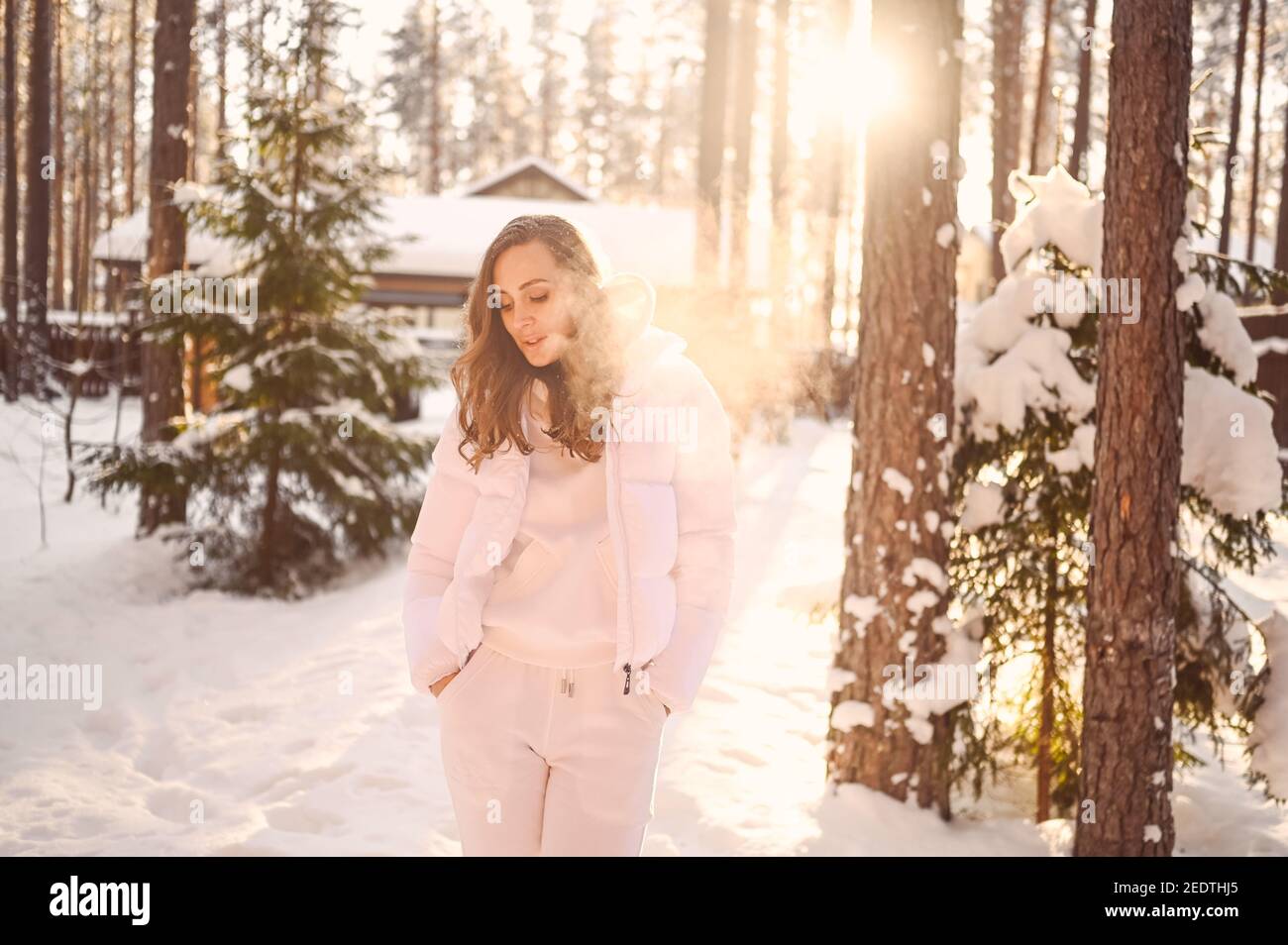 Hiver ensoleillé portrait froid dans la neige de la belle jeune femme dans une veste d'extérieur blanche chaude en duvet dans la cour de maison de campagne en bois sur le fond de pin d'épicéa vert arbres couverts de neige Banque D'Images
