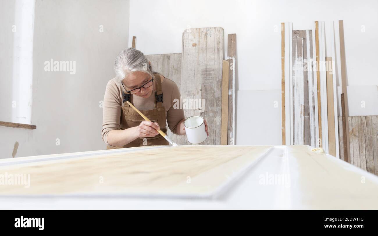 artisanat en bois, femme artisan charpentier peinture avec brosse et pot de peinture blanc la porte dans l'atelier, portant combinaison et lunettes, designer d'intérieur, Banque D'Images