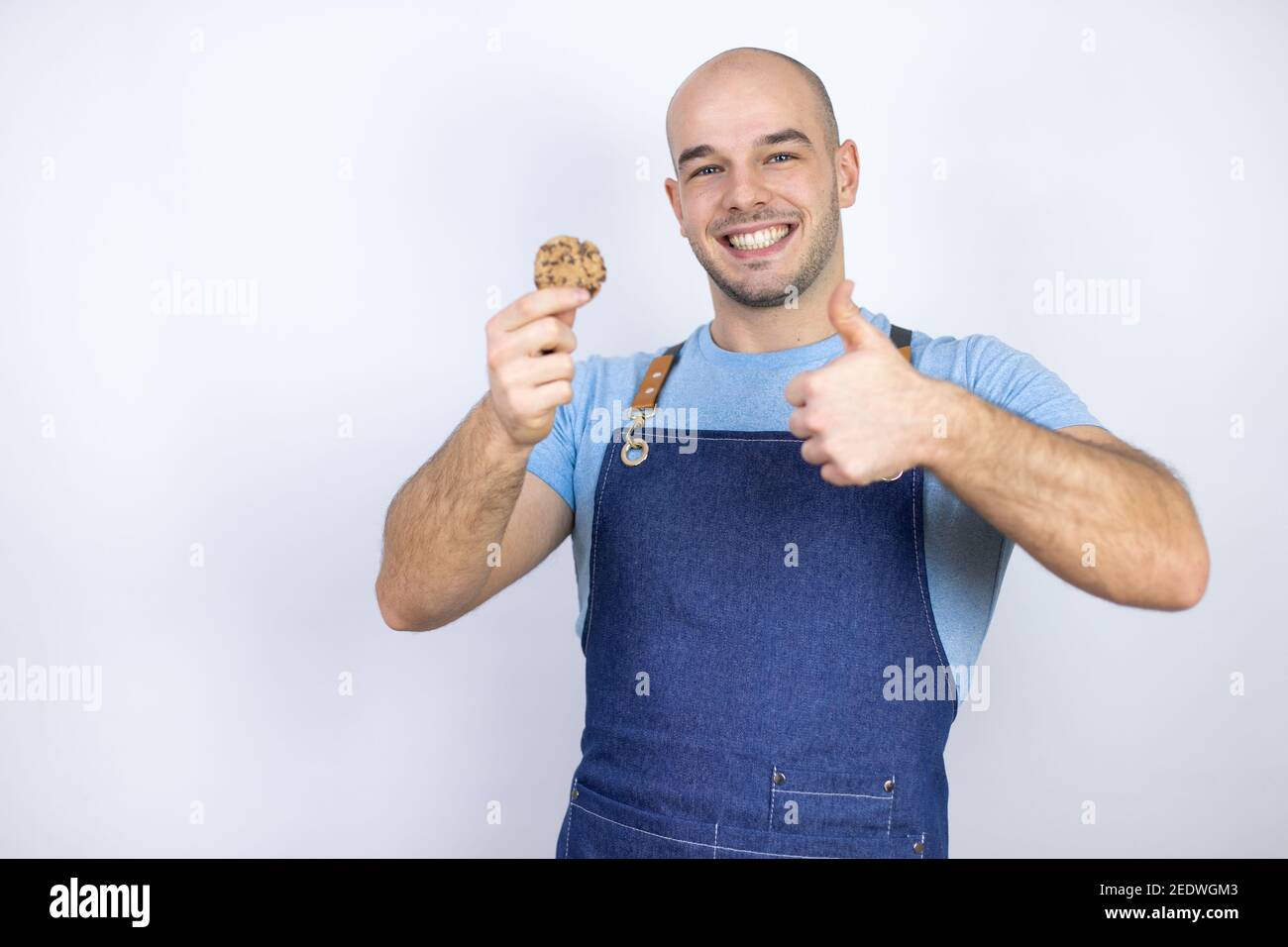 Jeune homme bald portant un tablier uniforme sur un fond blanc isolé Jeune homme bald portant un tablier uniforme sur un fond blanc isolé tenir un choc Banque D'Images