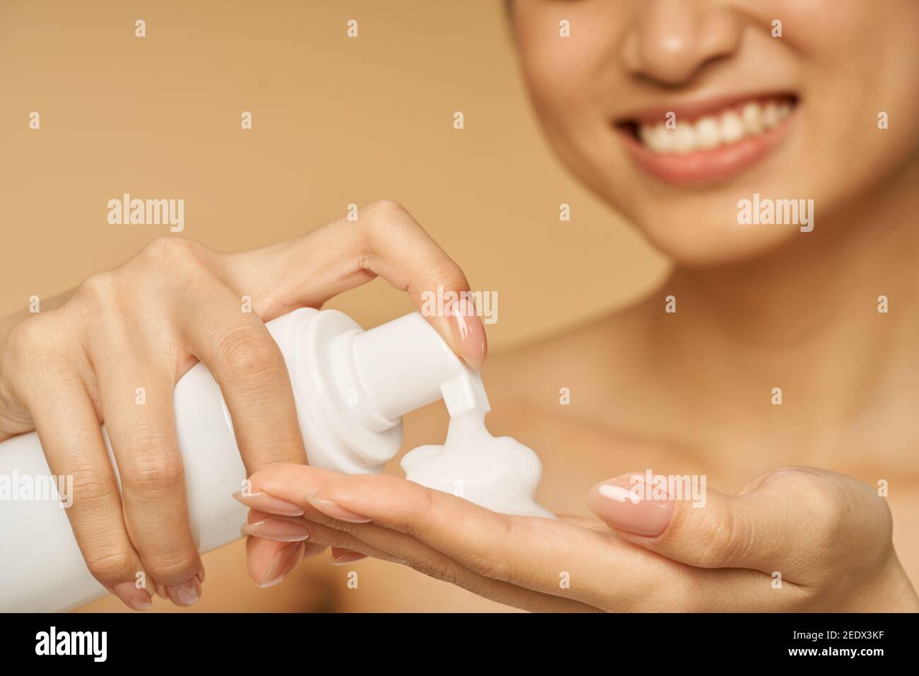 Prise de vue courte d'une jeune femme souriante, portant une bouteille de savon doux pour le visage en mousse isolée sur fond beige. Produits de beauté et concept de soins de la peau Banque D'Images