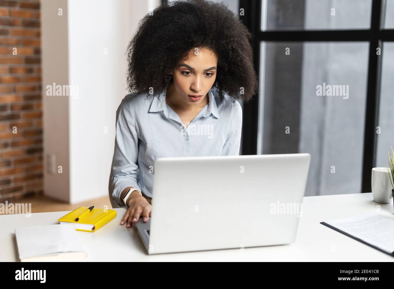 Jeune femme d'affaires de race mixte avec une coiffure afro assis à son bureau, a ouvert la bouche, en vérifiant les données du marché boursier en ligne, regardant Banque D'Images