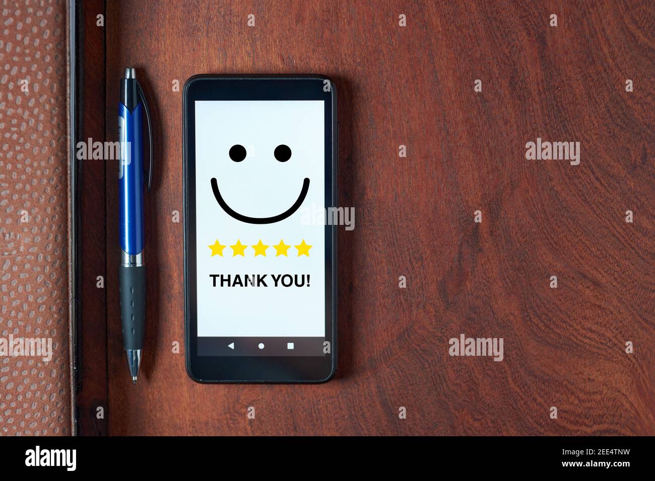 Vue de dessus du téléphone mobile avec visage souriant et note cinq étoiles avec le texte « Merci ! » sur un écran blanc, un stylo et un ordinateur portable posés sur un bureau. Banque D'Images