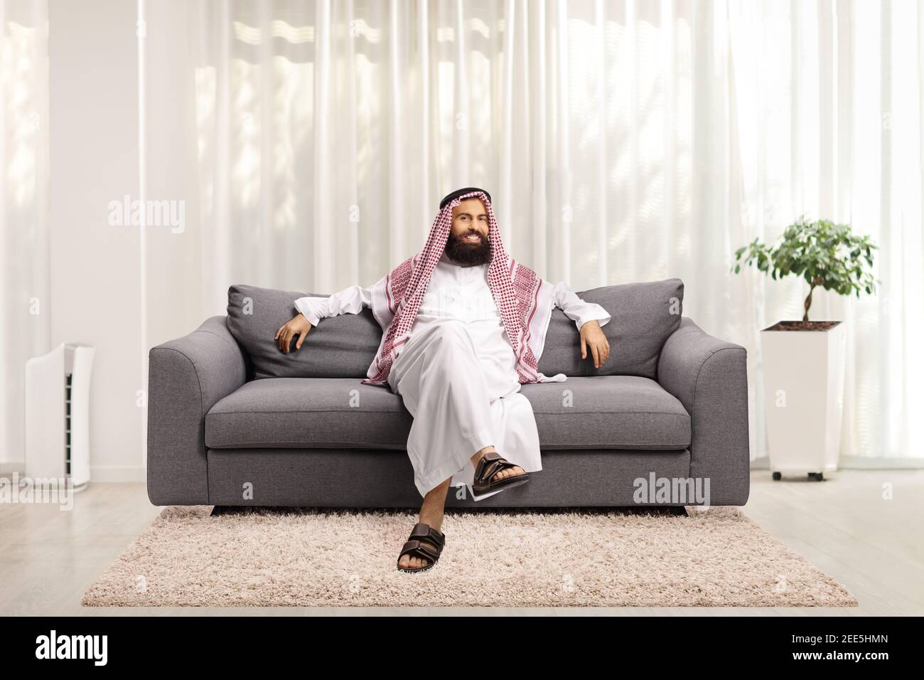 Homme arabe saoudien assis sur un canapé gris chez lui et regarder l'appareil photo Banque D'Images