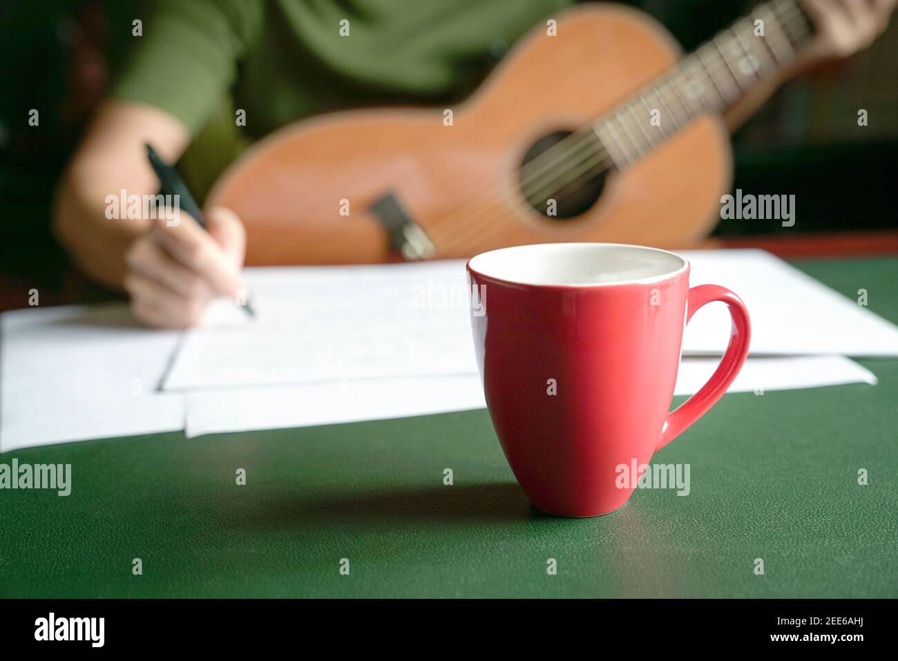 Blur femme musicienne avec guitare écrivant sur le livre au bureau. Mise au point sélective sur la tasse rouge. Banque D'Images