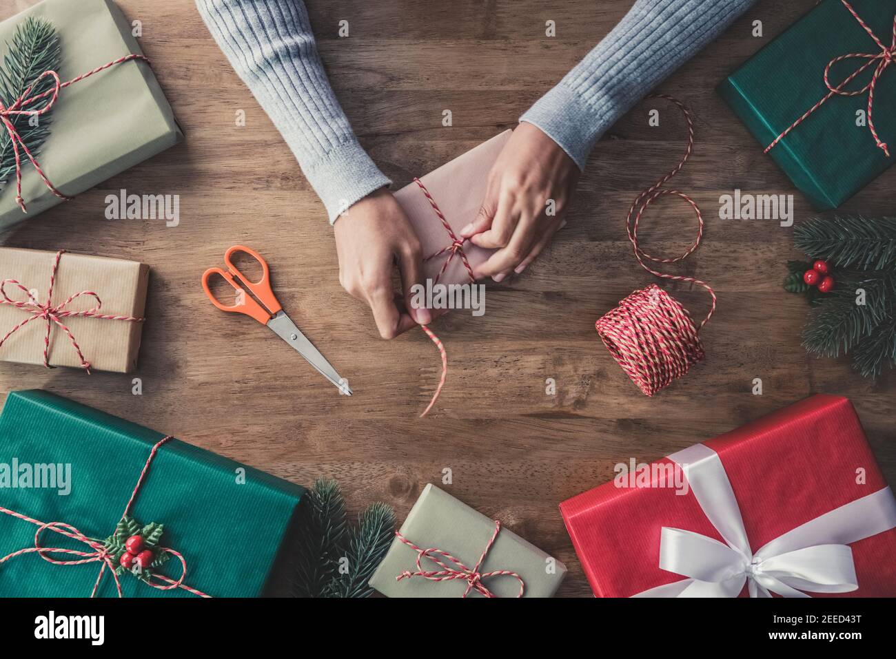 Femme mains dans un chandail gris emballage boîte cadeau avec l'équipement et des articles de décoration sur table en bois, se préparant pour célébrer les vacances de Noël - haut Banque D'Images