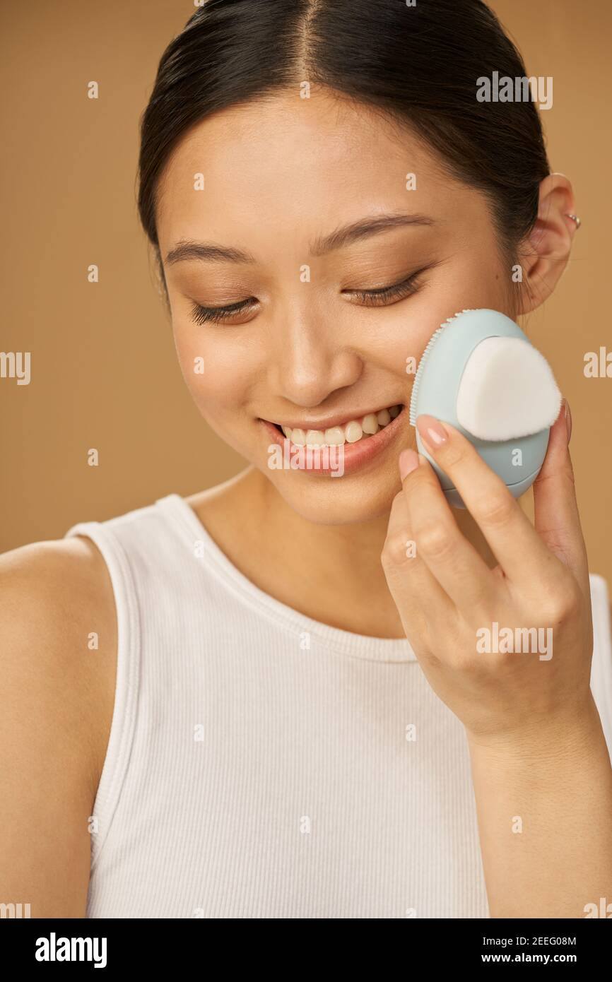 Jolie jeune femme souriant tout en utilisant la brosse de nettoyage facial en silicone, posant isolé sur fond beige. Accessoire de lavage. Concept de soin de la peau Banque D'Images