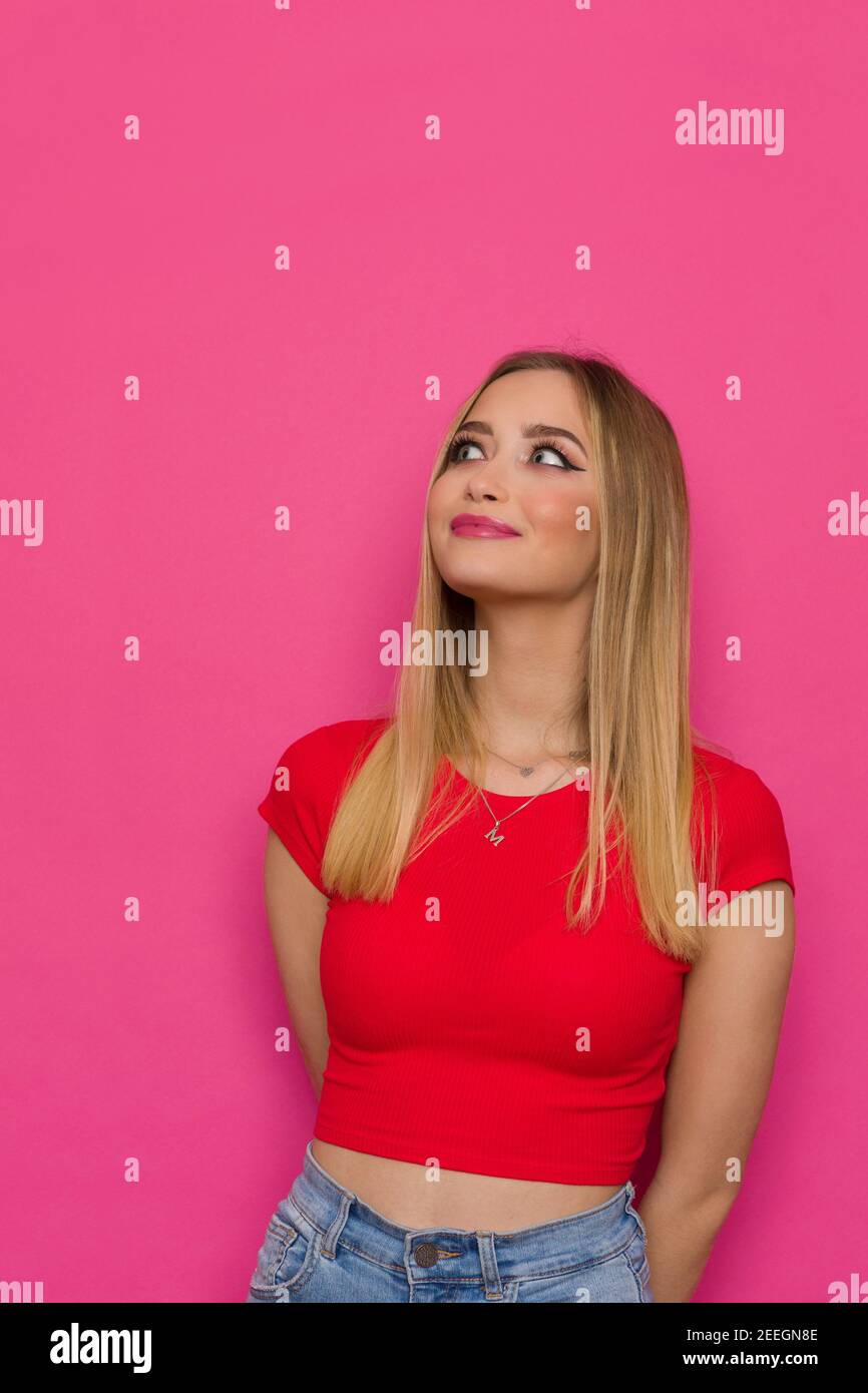 Curieuse jeune femme en haut rouge tient les mains derrière, regardant vers le haut et souriant. Taille haute studio tourné sur fond rose. Banque D'Images