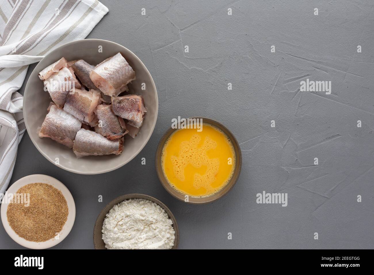 Dans un bol, des morceaux de merlu et des produits pour la cuisine frite, des œufs, de la farine et de la chapelure ont été présentés avec une serviette de cuisine sur fond gris Banque D'Images