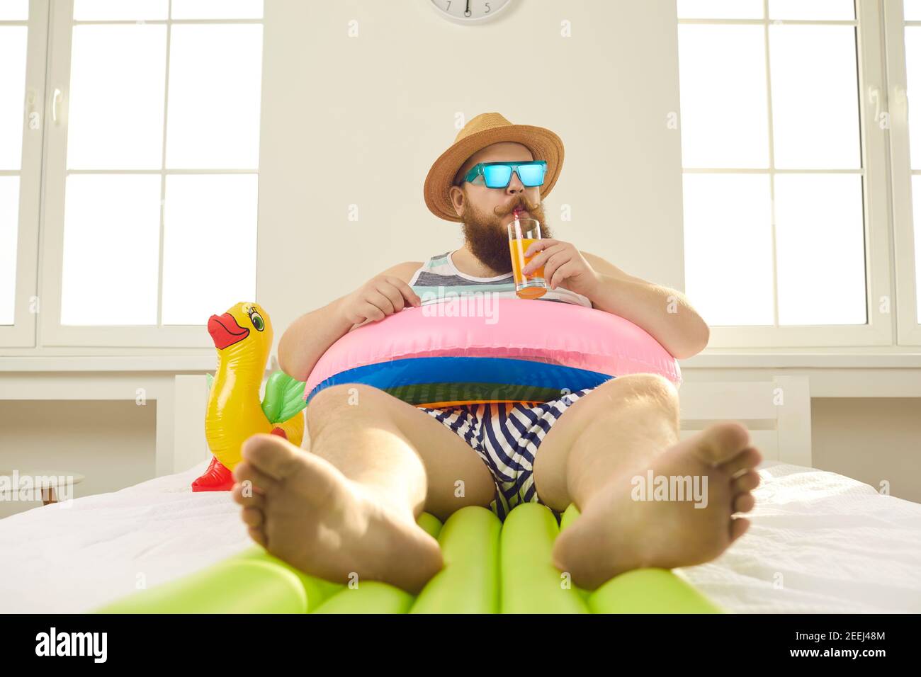 Drôle homme pieds nus en lunettes de soleil et vêtements d'été sirotant le jus en quarantaine à domicile Banque D'Images