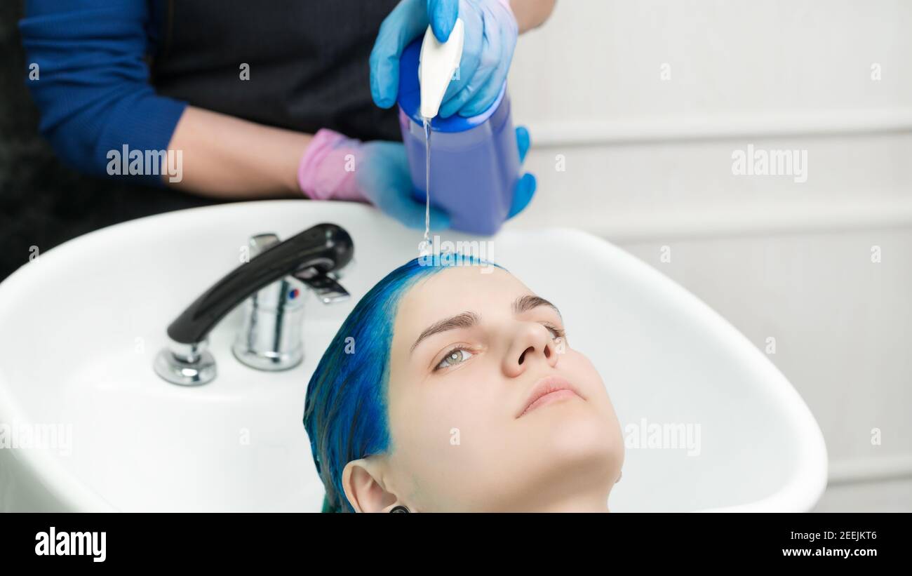 Le coiffeur passe les mains en gants pour comprimer le shampooing du tube dans la tête jeune femme avec des cheveux bleus tout en se lavant les cheveux en spécial salon de beauté avec lavabo et douche Banque D'Images