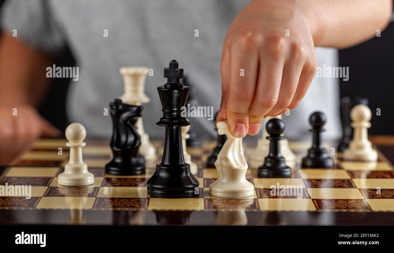 Un garçon caucasien tient une pièce d'échecs blanche au cours d'une partie. Il va faire un mouvement. Image d'arrière-plan sombre pour la détermination, la concentration, le foyer Banque D'Images