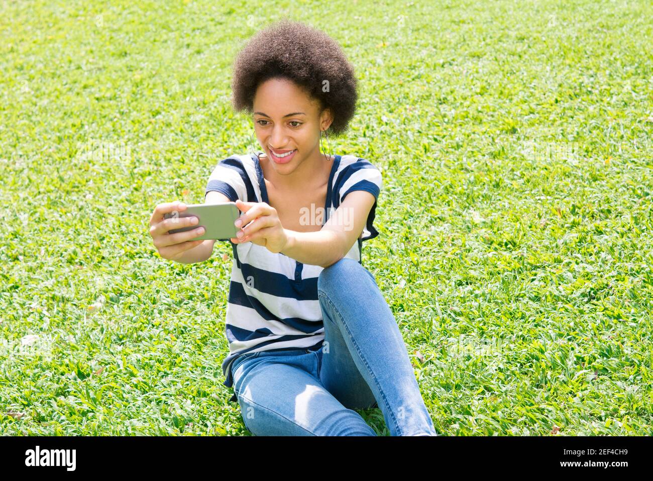 Jeune femme afro décontractée assise sur l'herbe prenant le selfie Banque D'Images