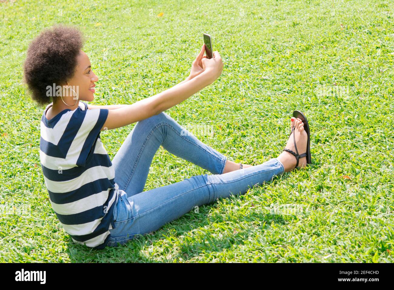 Jeune femme afro décontractée assise sur l'herbe prenant le selfie Banque D'Images