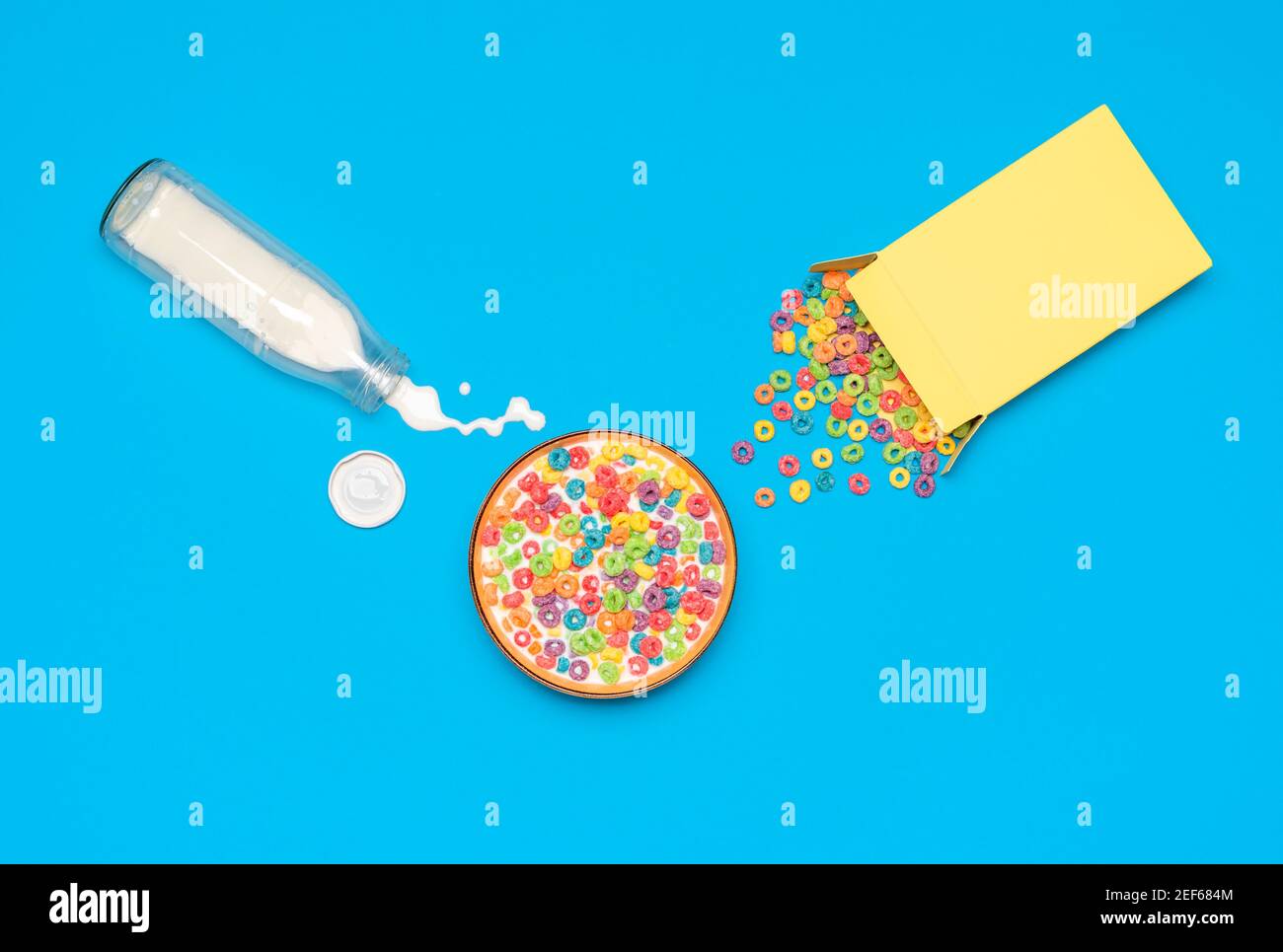 Vue ci-dessus avec des céréales colorées dans un bol et une bouteille de lait isolée sur un fond bleu. Table de petit déjeuner désordonnée avec du lait renversé et des céréales. Banque D'Images Vue ci-dessus avec des céréales colorées dans un bol et une bouteille de lait isolée sur un fond bleu. Table de petit déjeuner désordonnée avec du lait renversé et des céréales. Banque D'Images