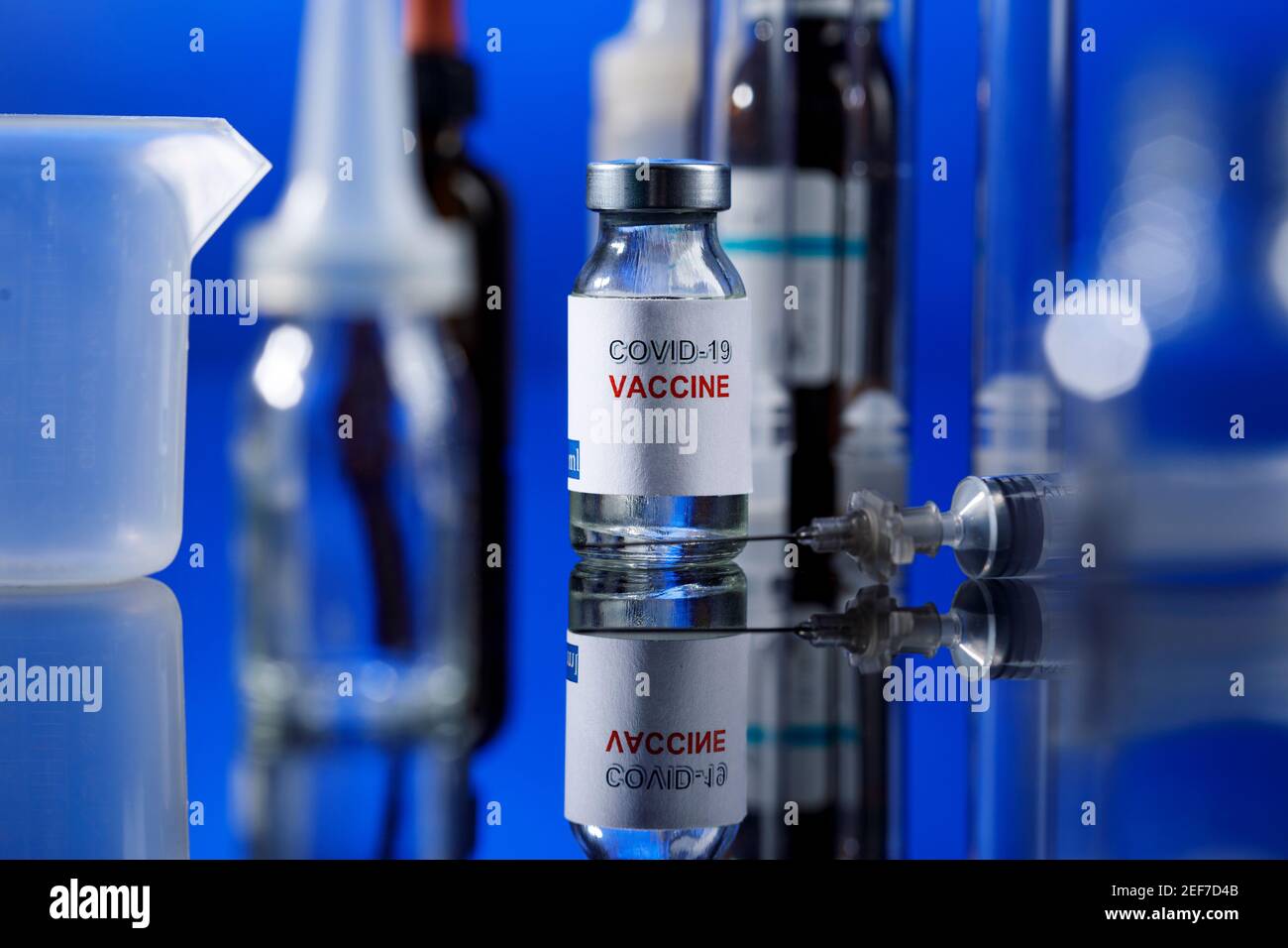 filet de verre de vaccin covid avec seringue et autres récipients de laboratoire placés sur une surface réfléchissante, isolés sur fond bleu Banque D'Images