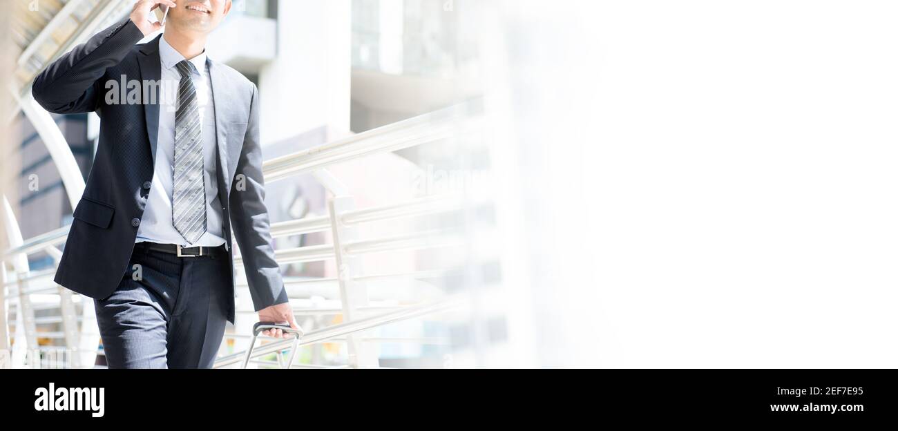 Businessman calling on cell phone tout en tirant des bagages et marche dans une allée couverte, vue panoramique sur copy space Banque D'Images