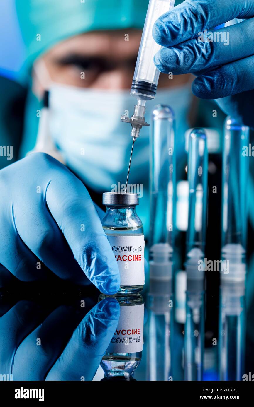 Médecin avec des capuchons médicaux et des gants en latex prend le vaccin covid avec une seringue dans un tableau avec plusieurs flacons Banque D'Images