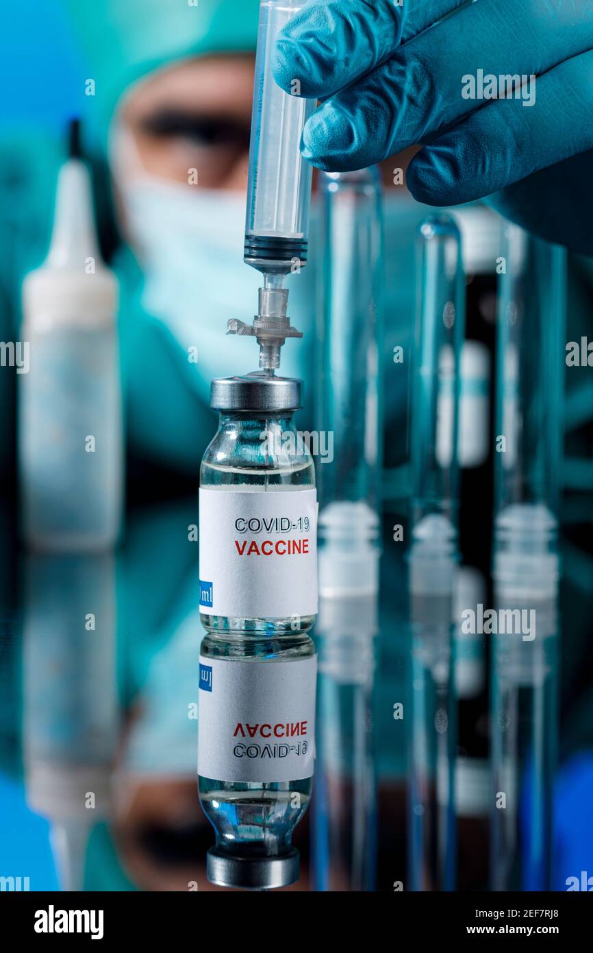 Médecin avec des capuchons médicaux et des gants en latex prend le vaccin covid avec une seringue dans un tableau avec plusieurs flacons Banque D'Images