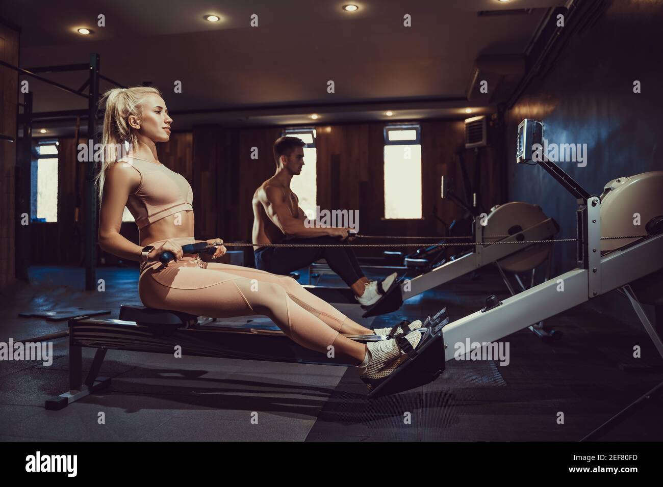 Vue latérale de la bonne fille blonde avec corps parfait dans l'entraînement de sport dos assis sur l'appareil d'exercice dans la salle de gym sombre atmosphère. Banque D'Images