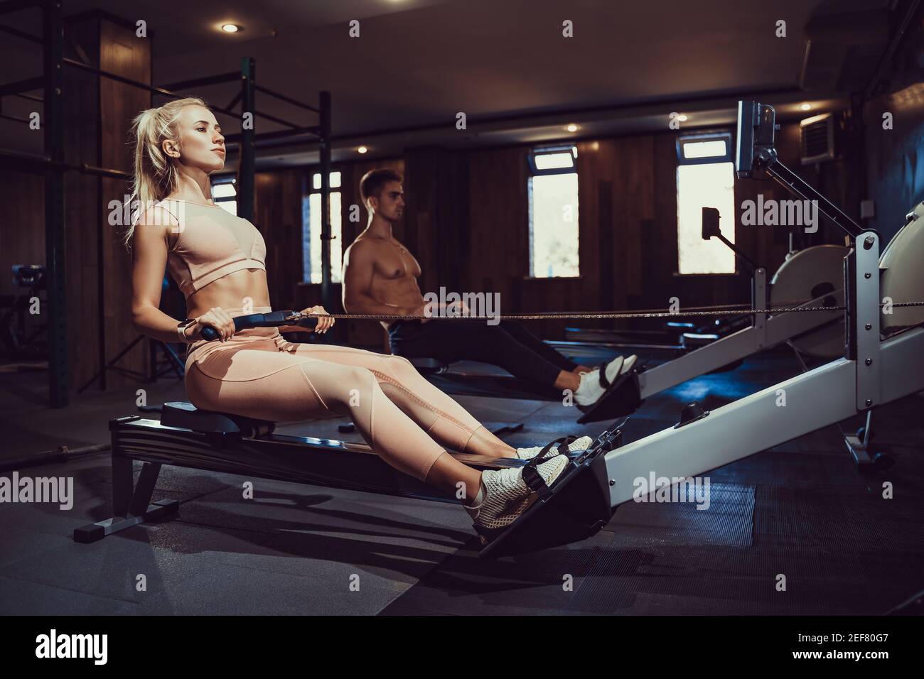Vue latérale de la bonne fille blonde avec corps parfait dans l'entraînement de sport dos assis sur l'appareil d'exercice dans la salle de gym sombre atmosphère. Banque D'Images