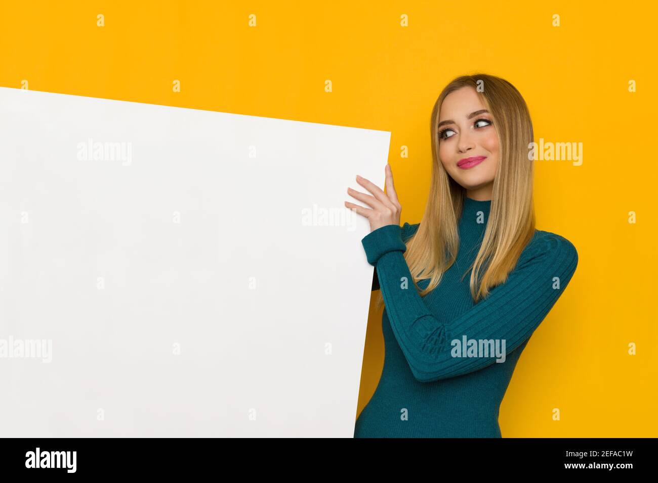 Jolie jeune femme souriante tient un écriteau blanc et regarde-le. Taille haute studio tourné sur fond jaune. Banque D'Images