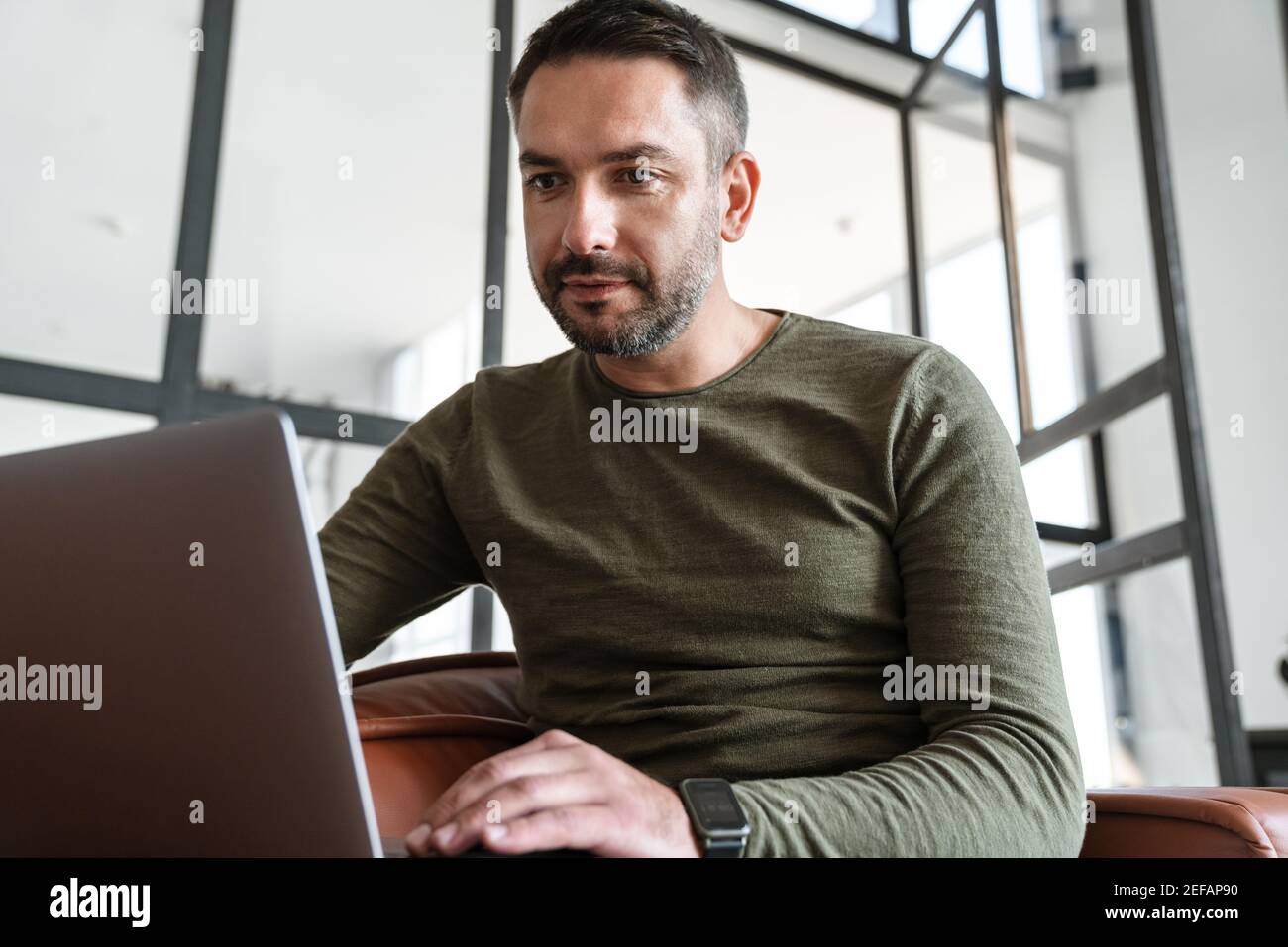 Beau jeune homme d'affaires souriant travaillant dans un espace de bureau moderne, utilisant un ordinateur portable, assis sur un canapé Banque D'Images