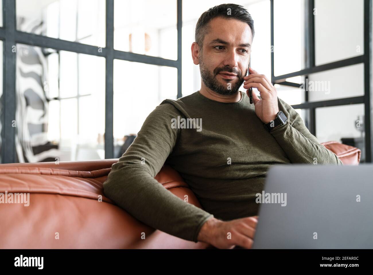 Beau jeune homme d'affaires confiant travaillant dans un espace de bureau moderne, utilisant un ordinateur portable, assis sur un canapé, parlant sur un téléphone portable Banque D'Images