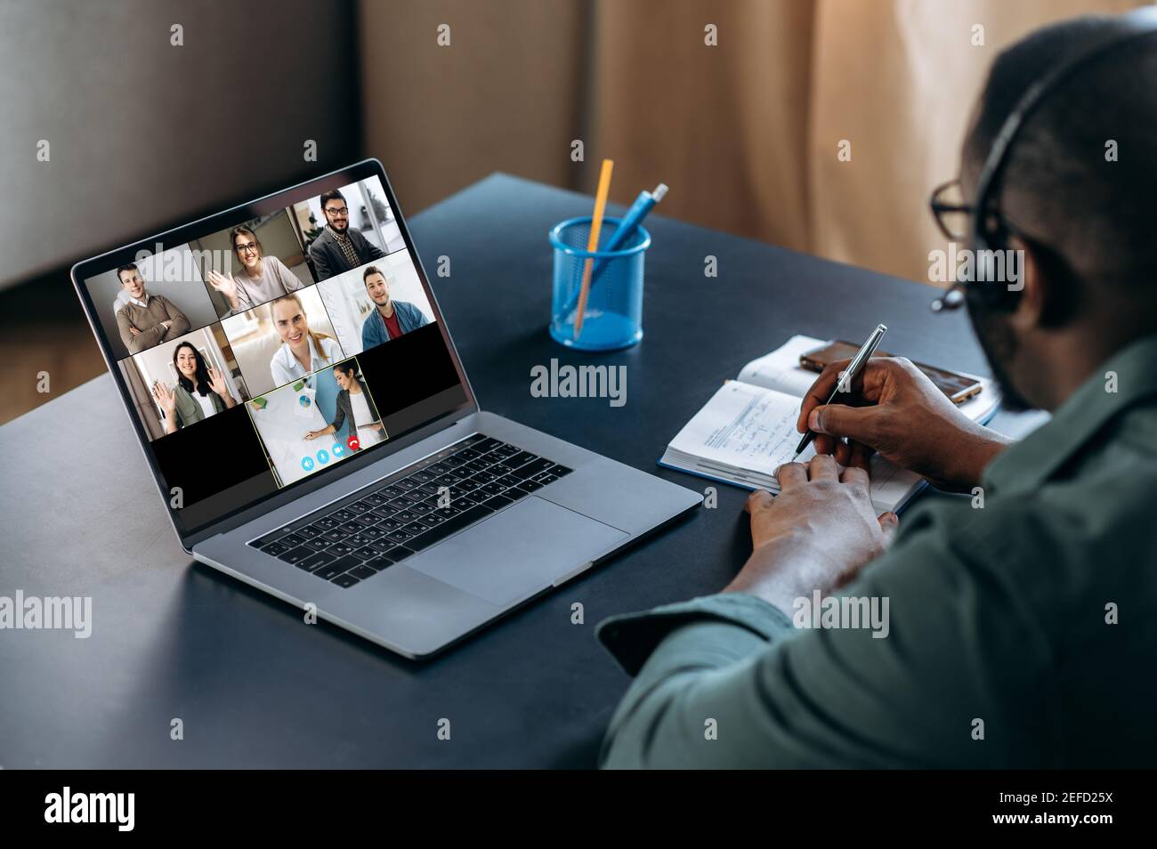 Enseignement à distance, cours en ligne. Un homme afro-américain dans un micro-casque apprend à distance et prend des notes à l'aide d'un ordinateur portable et d'une application de chat vidéo. Sur l'écran de l'ordinateur portable le professeur et le groupe de personnes diverses Banque D'Images