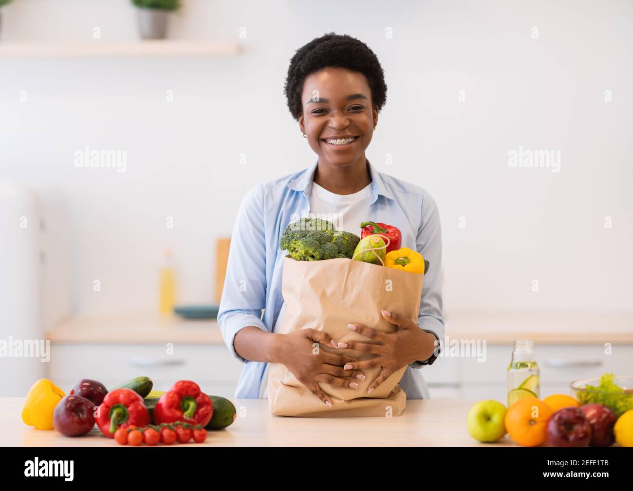 Joyeuse femme africaine tenant un sac d'épicerie plein de légumes biologiques et de produits alimentaires sains debout dans la cuisine à la maison, souriant à Camer Banque D'Images